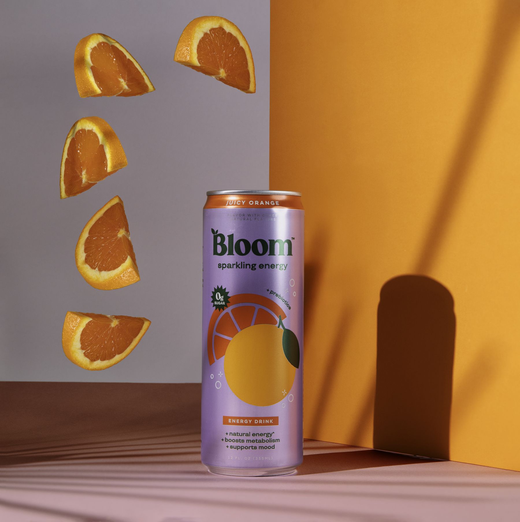 Bloom sparkling energy can (orange) with floating orange slices and a hard arch shadow on an orange/grey set.