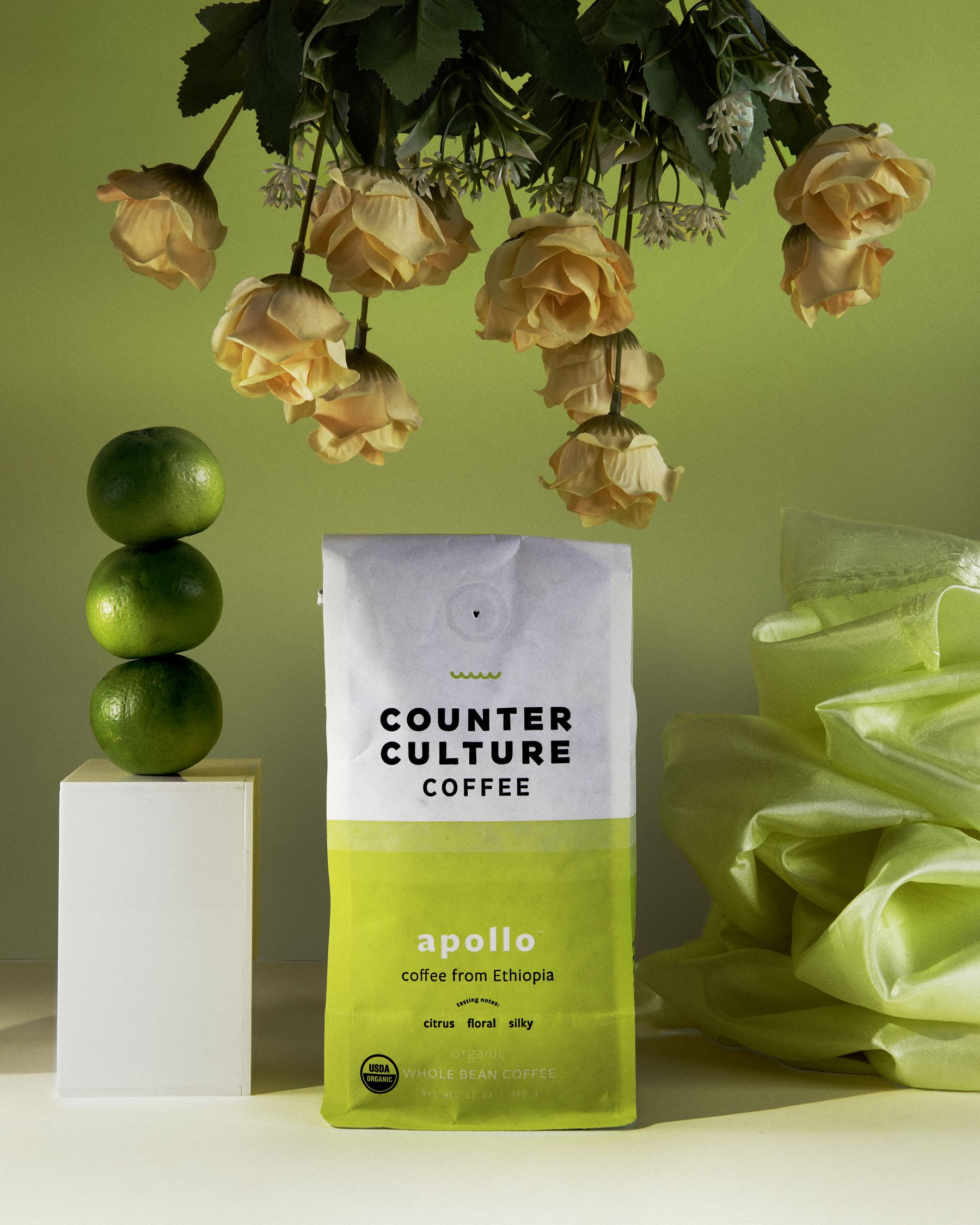 Counter Culture Coffee 'Apollo' bag with stacked limes on a plinth, hanging dried roses and chartreuse fabric.