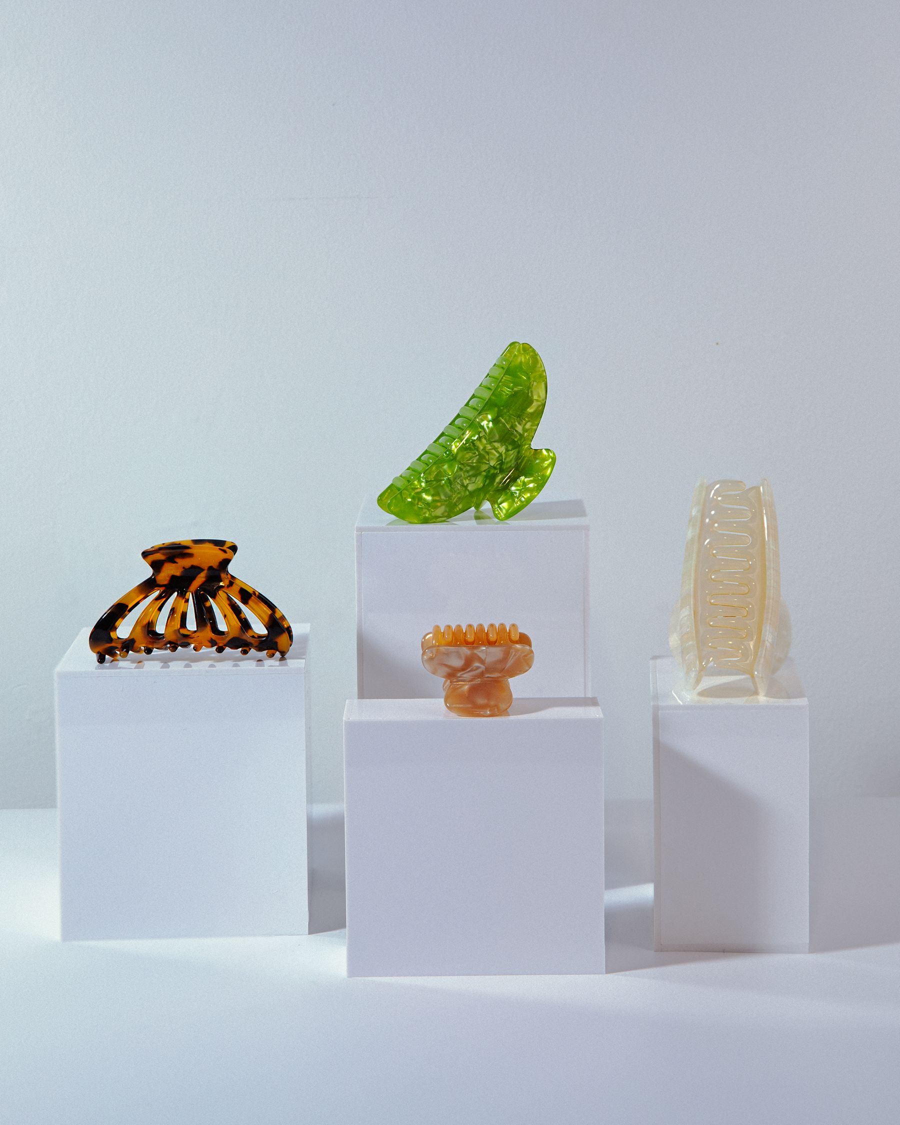Hair claw clips (tortoiseshell, green, clear) on white plinths.