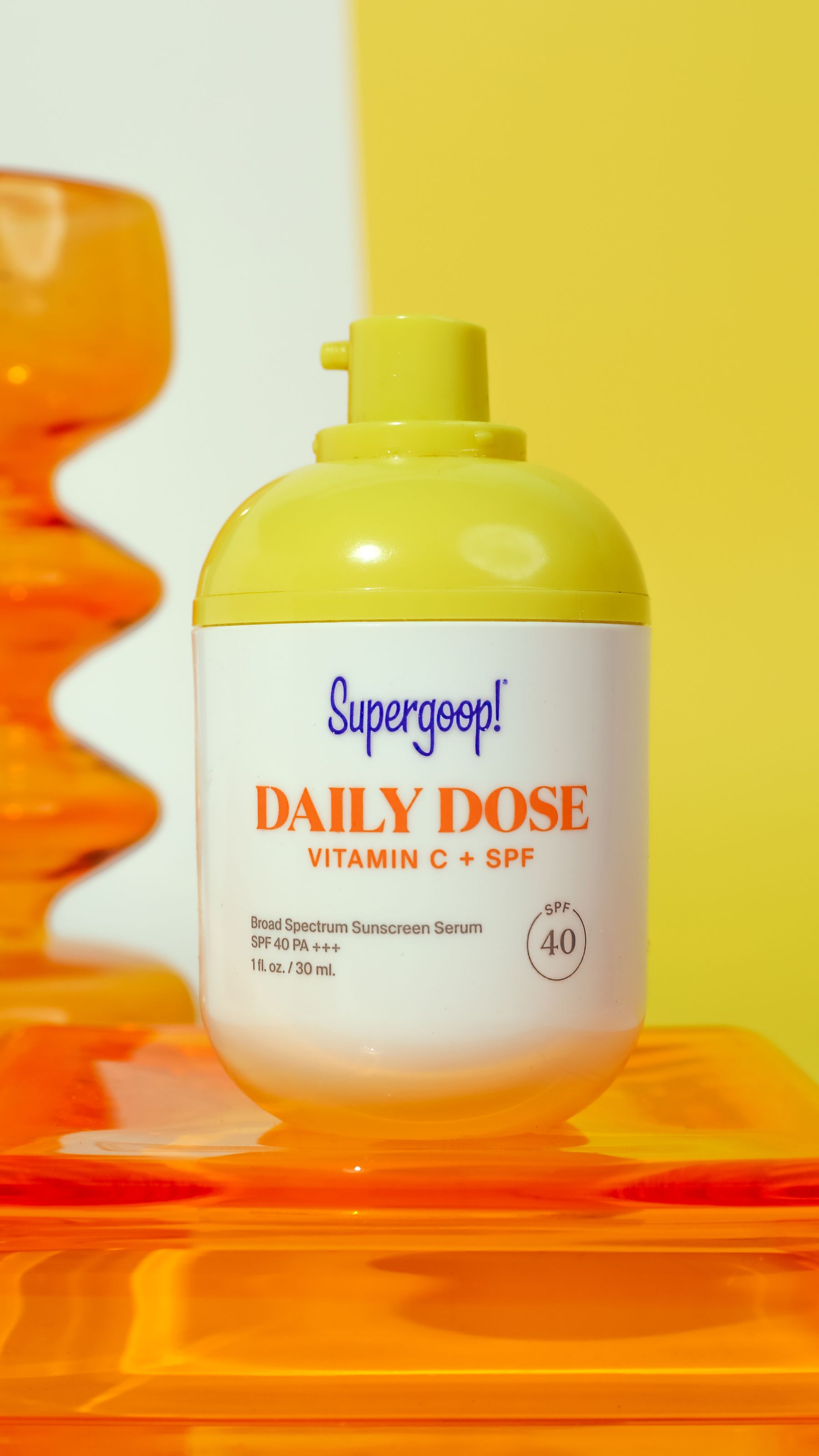 Supergoop! Daily Dose bottle with orange glass forms on a yellow/orange set (tall vertical).