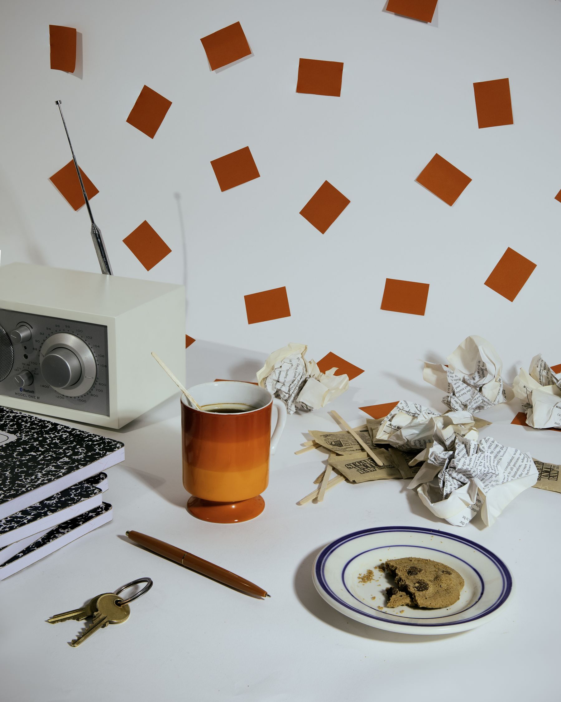 Desk still life: vintage radio, amber glass mug, notebooks, keys, pen and stained plate, wall of red-orange paper squares.