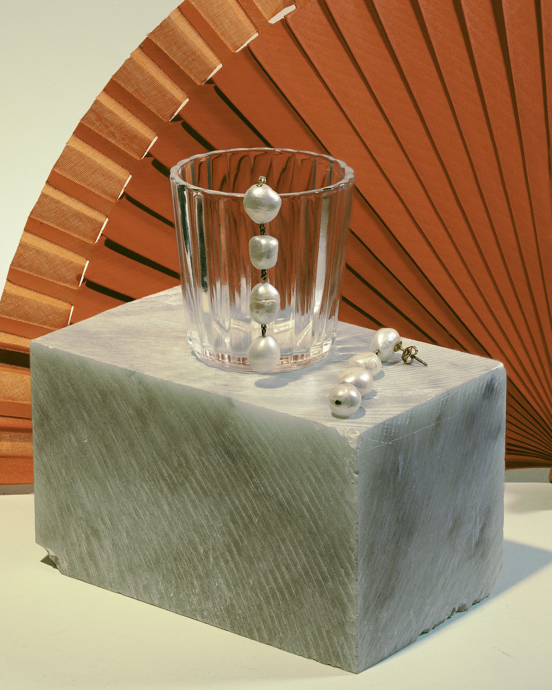 Glass tumbler with pearl strands on a marble block, wooden fan behind.