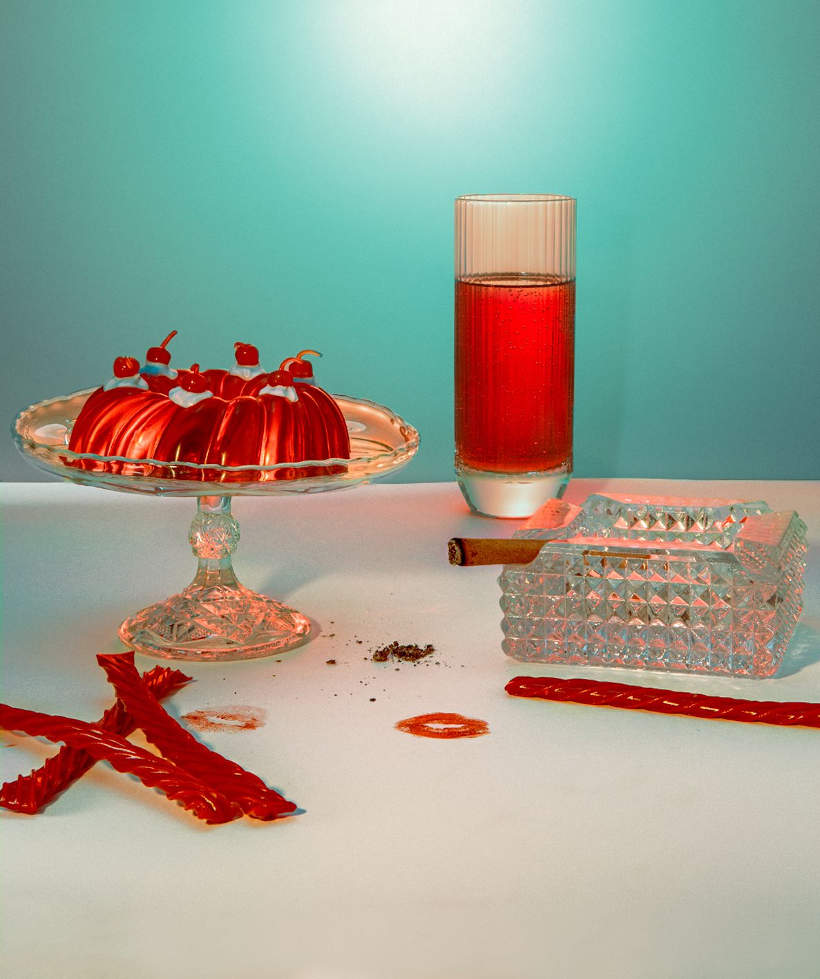Red jelly dessert on a glass cake stand with a red drink and red licorice on teal.